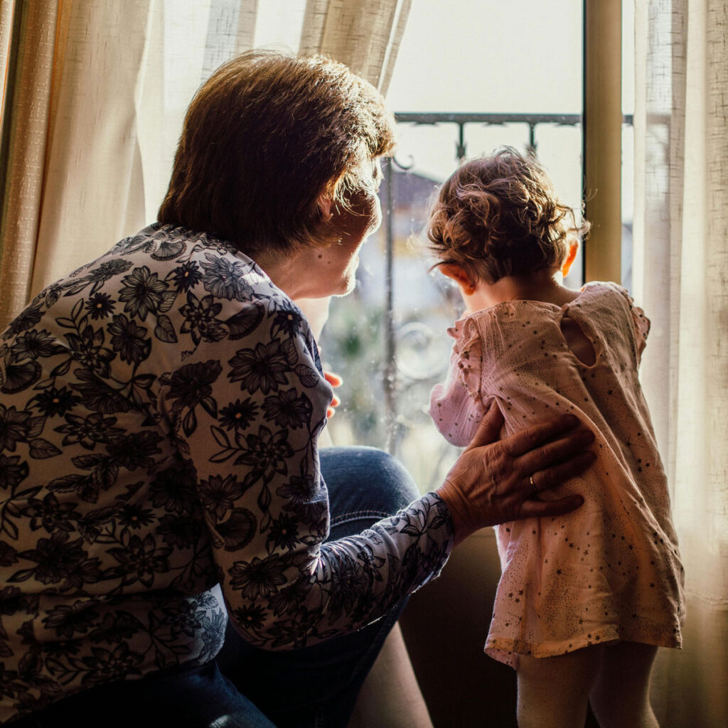 An older person and a baby looking out the window together in a cozy apartment.